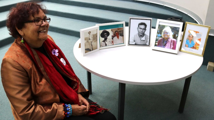 Marlene Jackamarra sits in a chair with pictures of her relatives sitting on a table beside her.