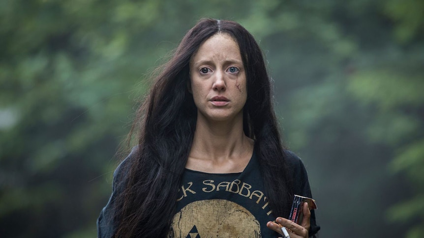 Colour mid-shot still of a stricken Andrea Riseborough walking in the woods holding a book and cigarette in 2018 film Mandy.