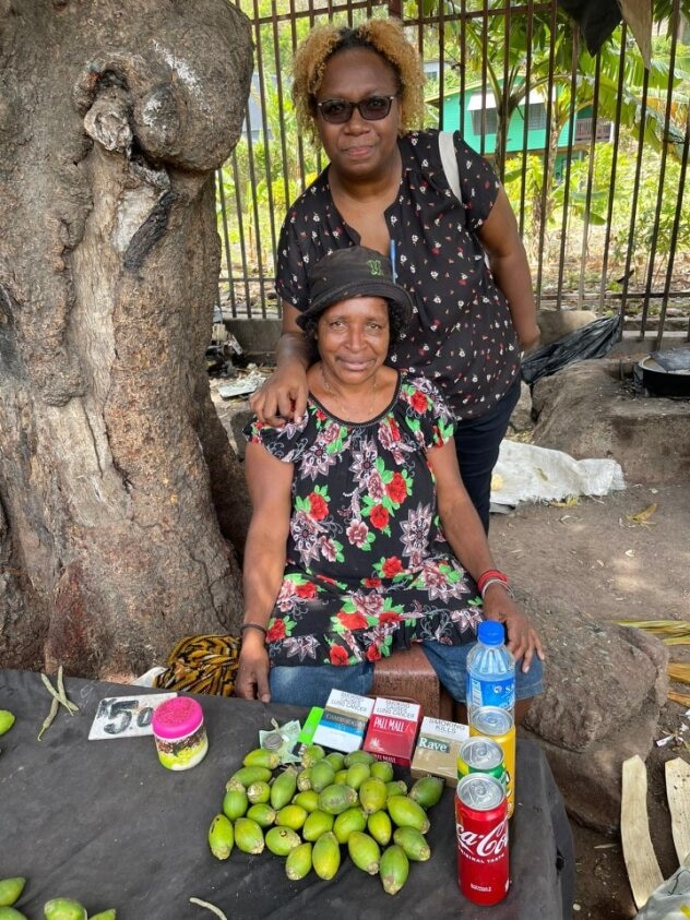 Kobieta stoi za kobietą sprzedającą powai, siedząc przy stole obok zielonych owoców, takich jak orzechy betelu. 