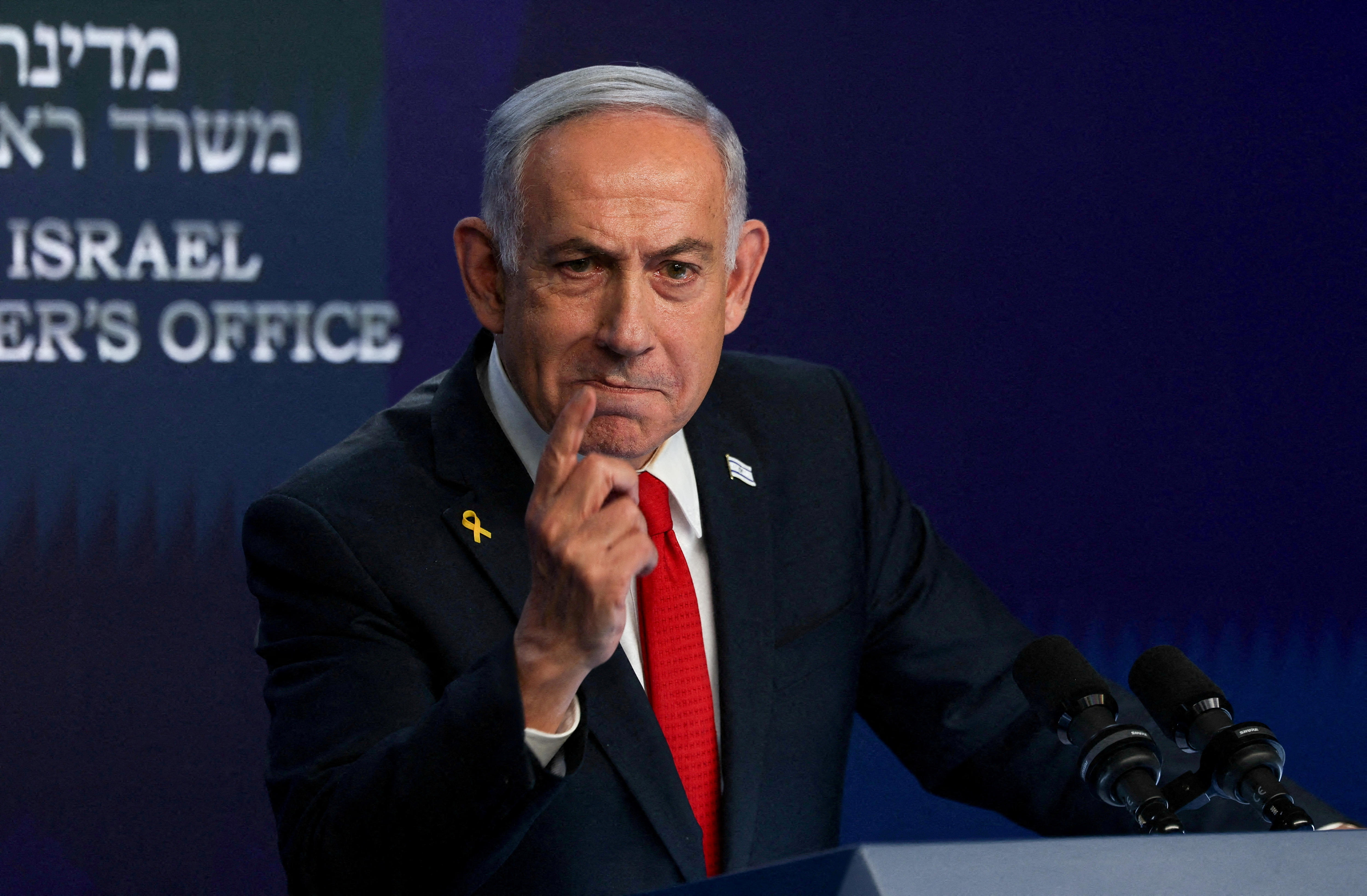Netanyahu, wearing a yellow ribbon pin and an Israeli flag pin, raises a finger and looks serious.