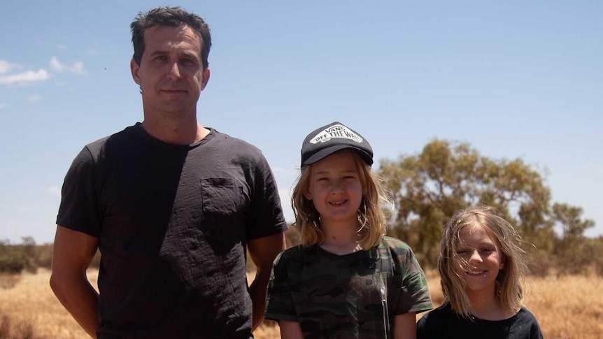 A man and his two daughters stand on scrubby bushland.