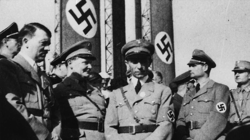 阿道夫·希特勒（左）与一些高级纳粹分子站在前面纳粹旗帜。” class=