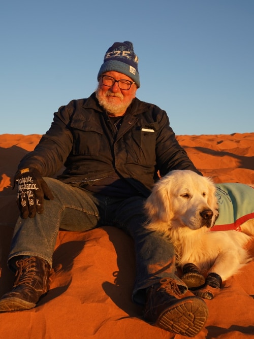 A man and a dog sit together on a red sand dune in the early morning.