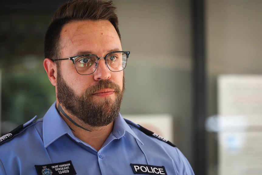 A male police officer with tattoos and glasses stands in uniform looking stern away from the camera.