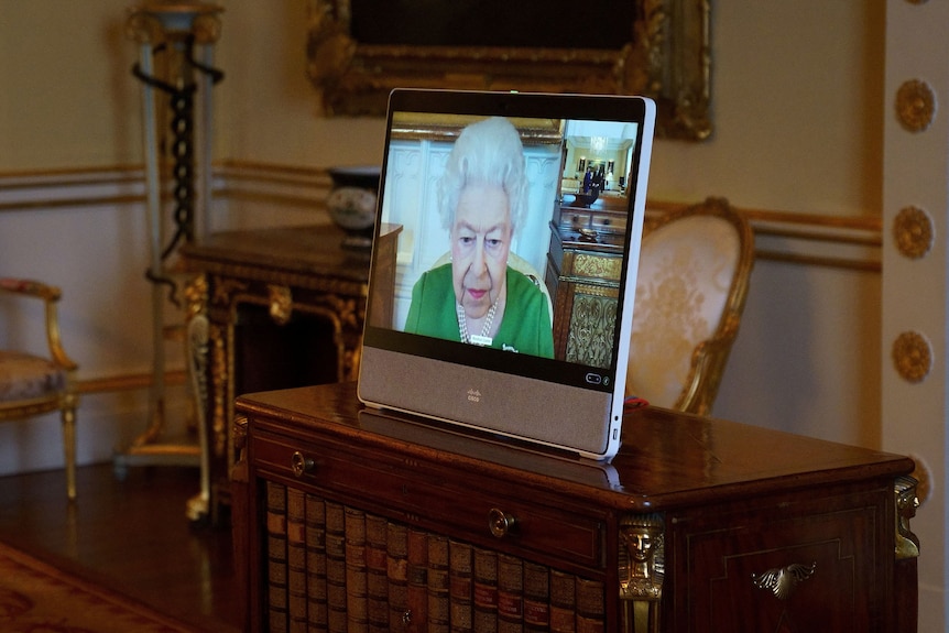 Una pantalla con la reina Isabel aparece sobre un escritorio de madera en una majestuosa sala del Palacio de Buckingham.