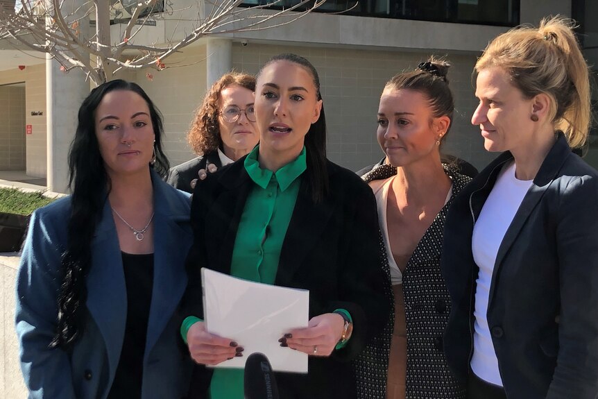 Four women stand together, with a lawyer behind, as one reads from a piece of paper.