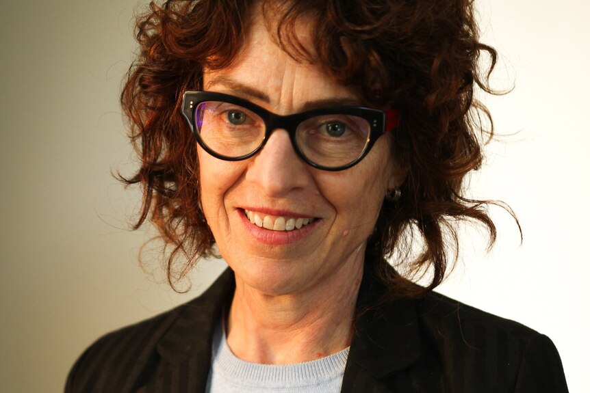 A middle aged woman with mid-length dark red hair and black-rimmed spectacles smiles posing for a photo.