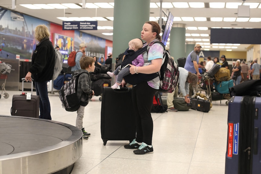 A young woman with a little boy and a baby strapped to her chest stands at baggage claim