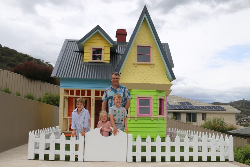 A man stands with his three children in the front yard of a massive cubby house.