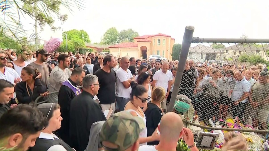 a crowd praying next to a broken fence on a suburban street