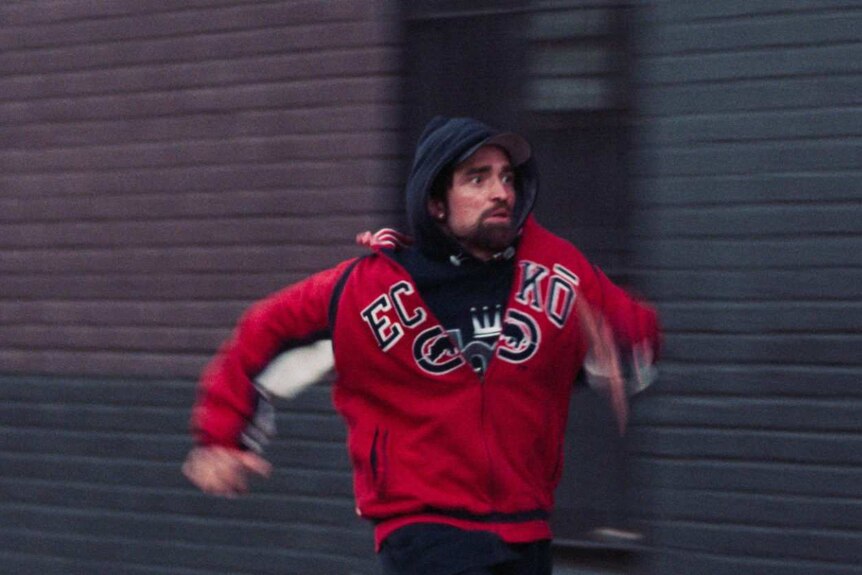 Still colour image from 2017 film Good Time of Robert Pattinson running through a street scene.