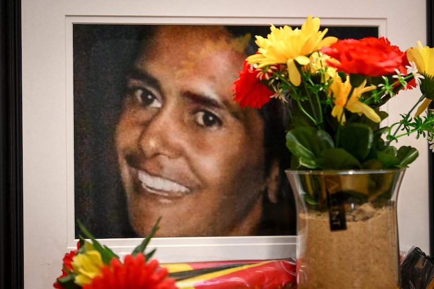 A photo of a woman's smiling face next to a vase of bright-coloured flowers.