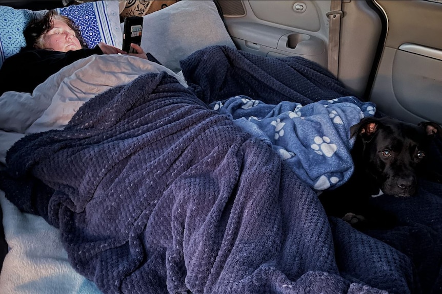 Mandy sleeps under a doona in the back of her car. A black dog sits on the end of her doona.