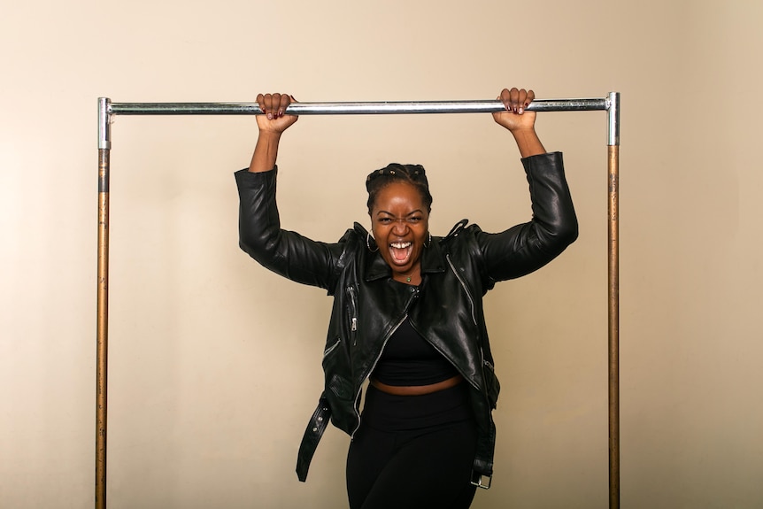 Ngwenya, a Black woman with braided hair, wears a black leather jacket and hoop earrings leans on a silver clothes rack, beaming
