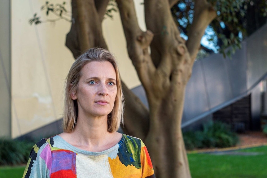 Gabrielle Appelby is looking into the distance, with a Morton bay fig behind her. She is blonde with blue eyes.