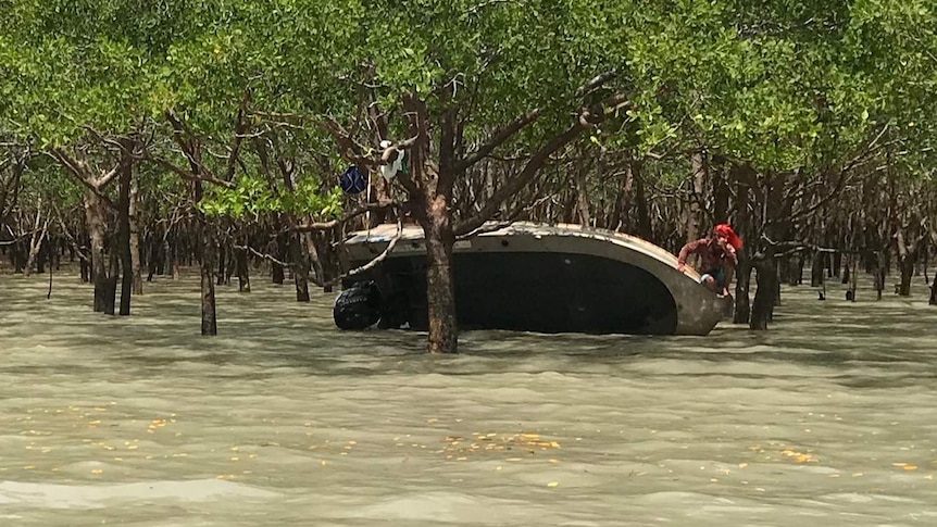 The boat is pictured on its side, near mangroves. A man clings to a tree.