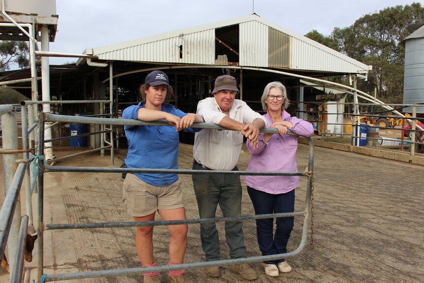 A middle-aged man and woman stand at a farm gate lean on a farm gate at a dairy with a younger woman