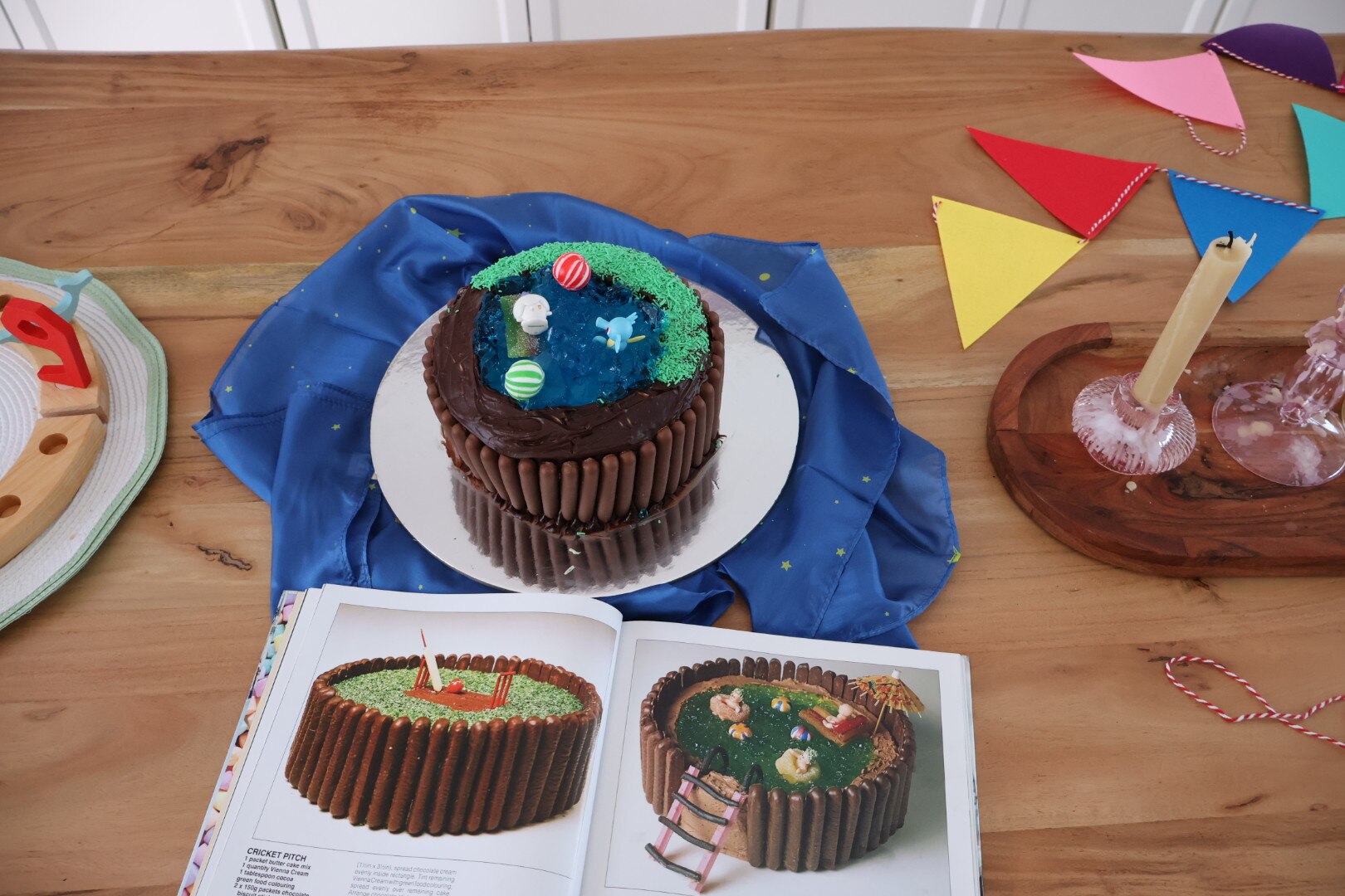 A chocolate cake with a jelly pool on its top with animal figurines and candy balls. Below is an open Woman's Weekly cake book.