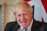 Boris Johnson grimaces while sitting behind a Union Jack flag 