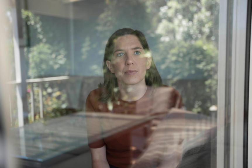 a woman sits by a window looking out, the reflections over her face