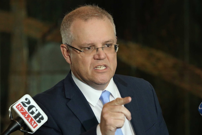 Scott Morrison bares his teeth and points a finger while talking. There are microphones around him.