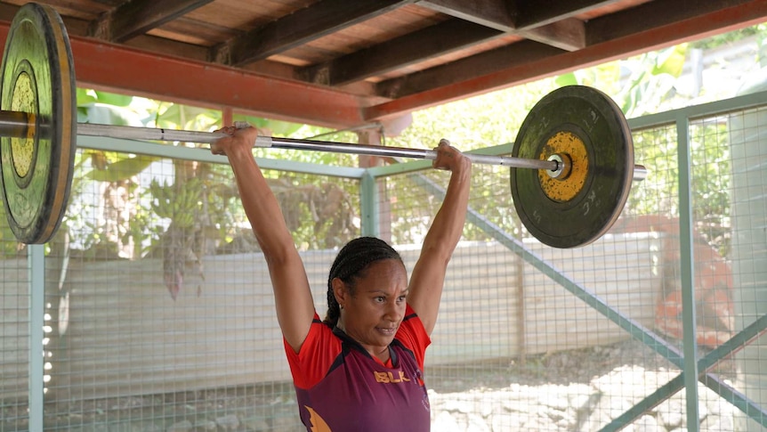A Black woman wearing a maroon shirt lifts a barbell loaded with weight plates.