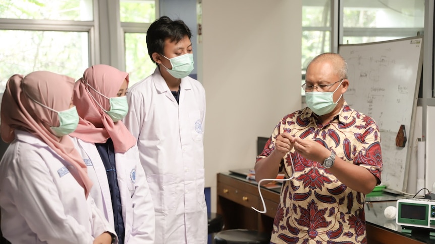 A man wearing a mask and glasses stares at a device in his hands as a man and two women wearing masks watch on.