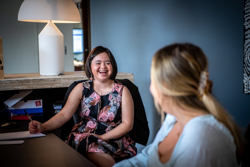 Olivia Sidhu sitting at a large table in an office, laughing and smiling with a blonde woman.