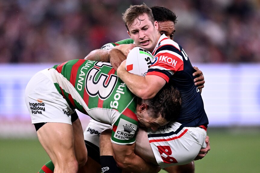 A Sydney Roosters NRL player holds the ball as he is tackled by two South Sydney opponents.