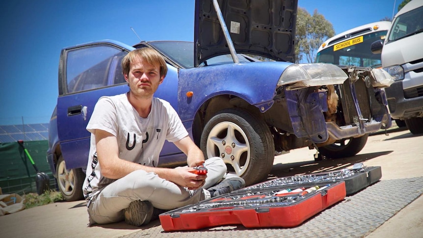 Markus sits on the ground beside the car he is rebuilding.