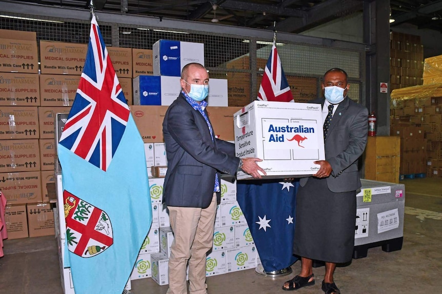 A man hands a box labelled Australian Aid to another man standing in front of a pile of boxes and Fijian and Australian flags.