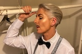 A drag king with a moustache in a suit and suspenders holds onto a pole inside a closet.