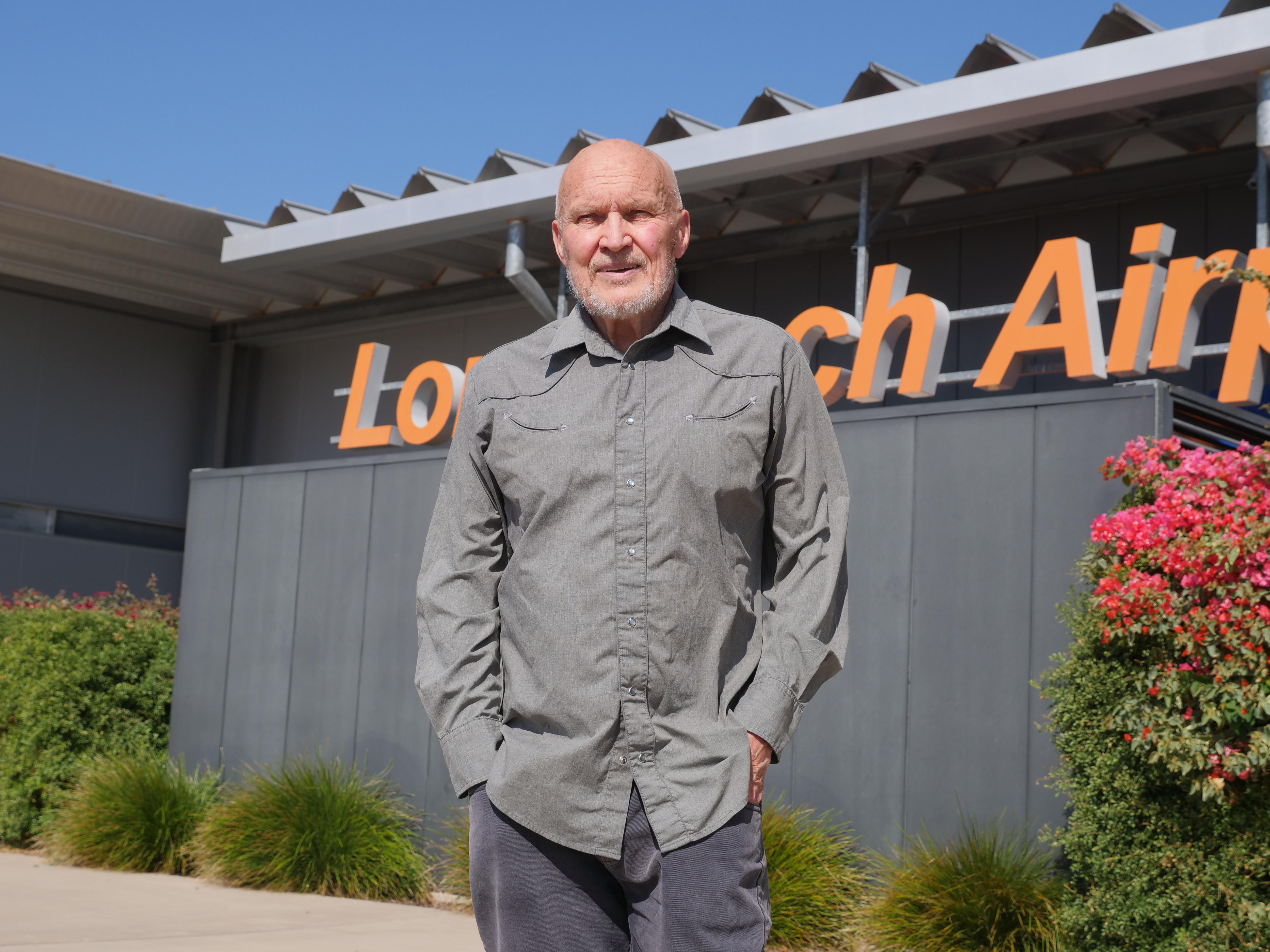 A man wearing a long-sleeved outback workshirt has a stern expression, standing in front of a "Longreach Airport" sign.