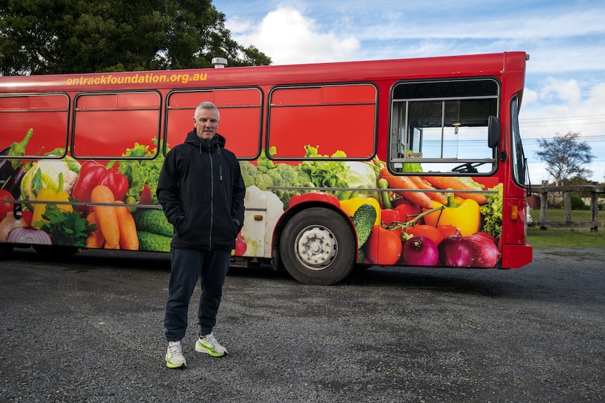 A man stands in front of a big bus with pictures of vegetables on the side of it.