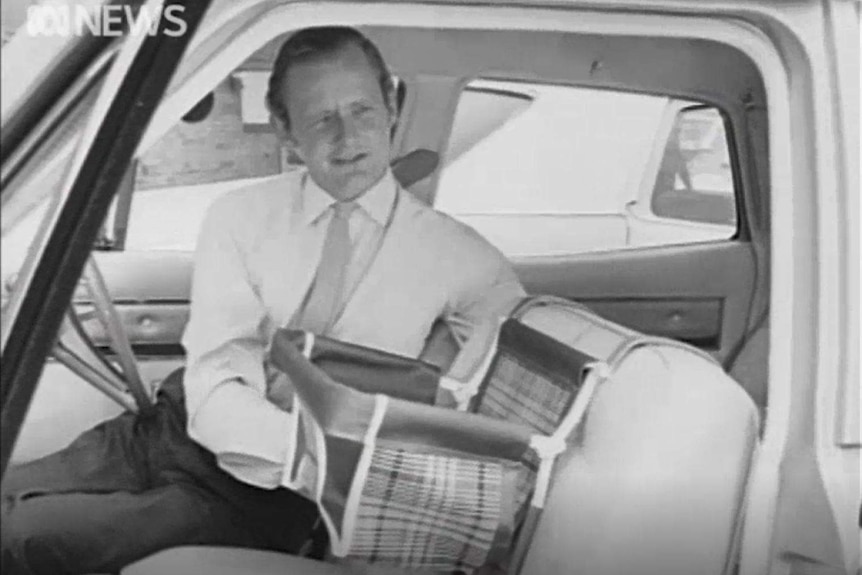 A still from a black and white video shows a man demonstrating a baby seat