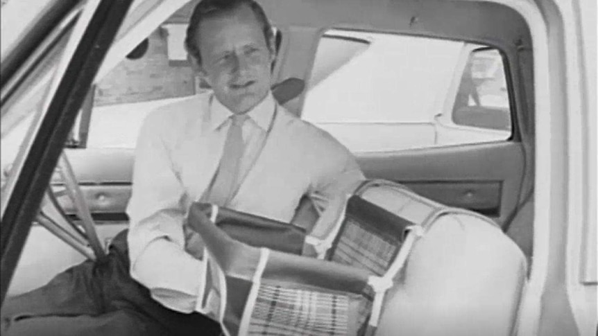 A still from a black and white video shows a man demonstrating a baby seat