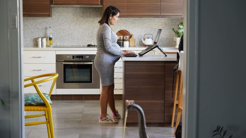 A pregnant woman in a grey dress stands at a kitchen bench working on a laptop.