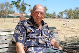An elderly man sitting on a bench in a cemetery, with graves behind him.