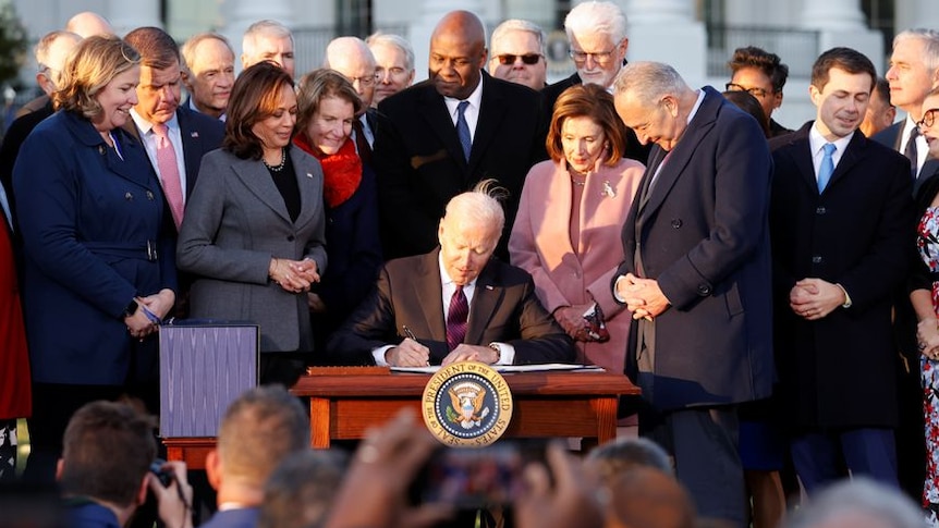 People in business suits and coats crowd around a man at a small table while he signs a document on a bright day