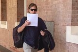 Man in blue button shirt with slacks, sunglasses and coat shields his face with a piece of paper as he walks into courthouse