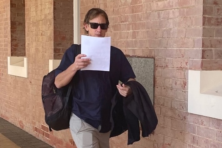 Man in blue button shirt with slacks, sunglasses and coat shields his face with a piece of paper as he walks into courthouse