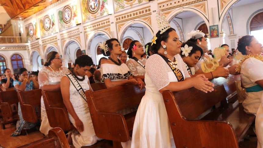 Pews of Samoan women in white wedding-style dresses are pictured kneeling and praying.