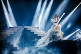 A young man stands on a staircase holding a microphone, wearing a white dress and a crystal mask surrounded by spotlights.