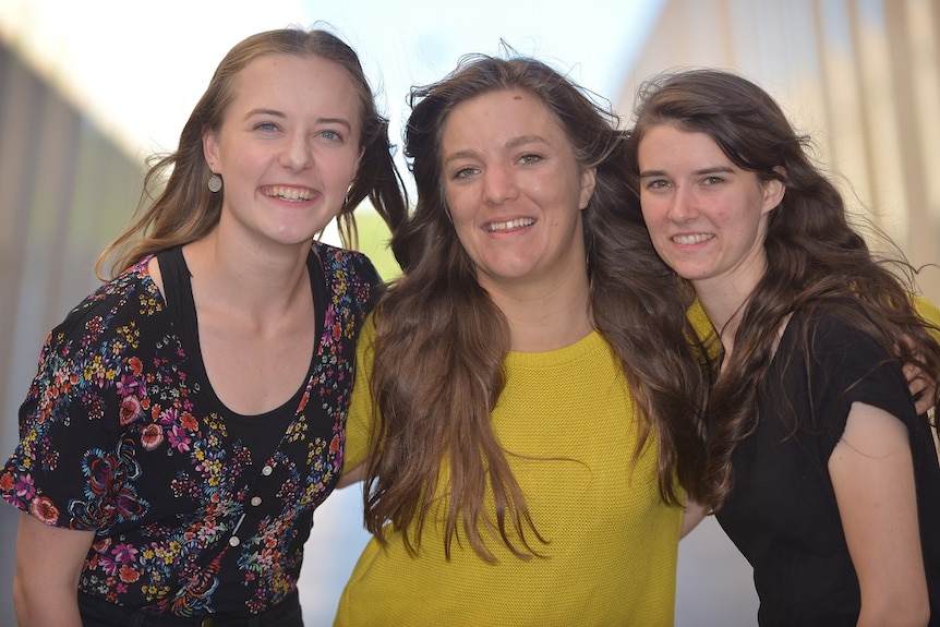 Three women smile at the camera with a blurred corridor behind them