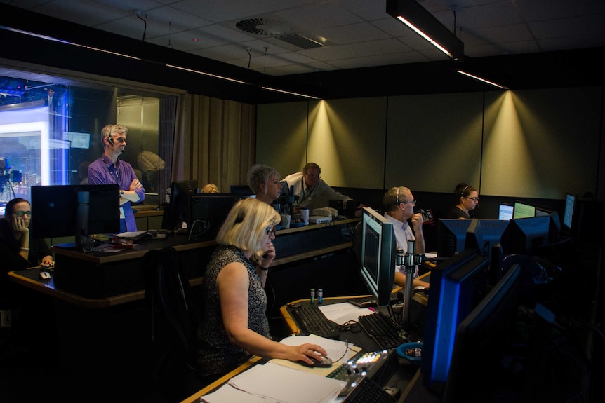 Eric Napper standing at back of control room with headphones on with production staff sitting in front of screens in front.