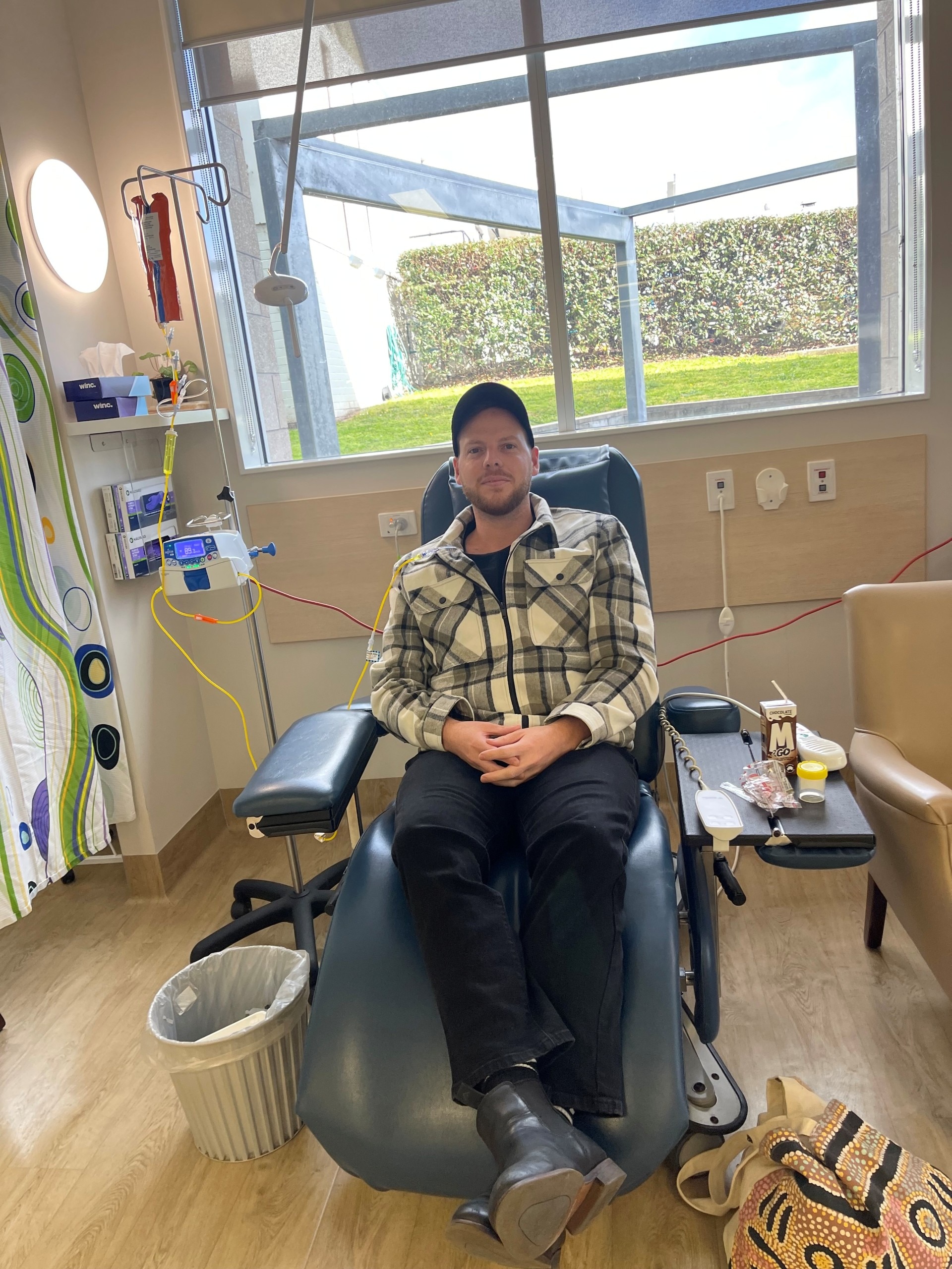 A young man wearing a cap sits in a chair getting chemo. 