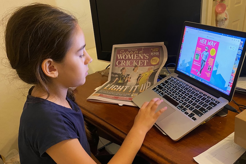 An 11-year-old sits in from of a laptop with a women's cricket newspaper liftout on the desk.