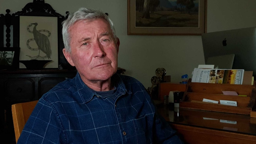 Bernard Collaery wearing a dark blue shirt with light blue stripes, sitting at a wooden desk in his front room office