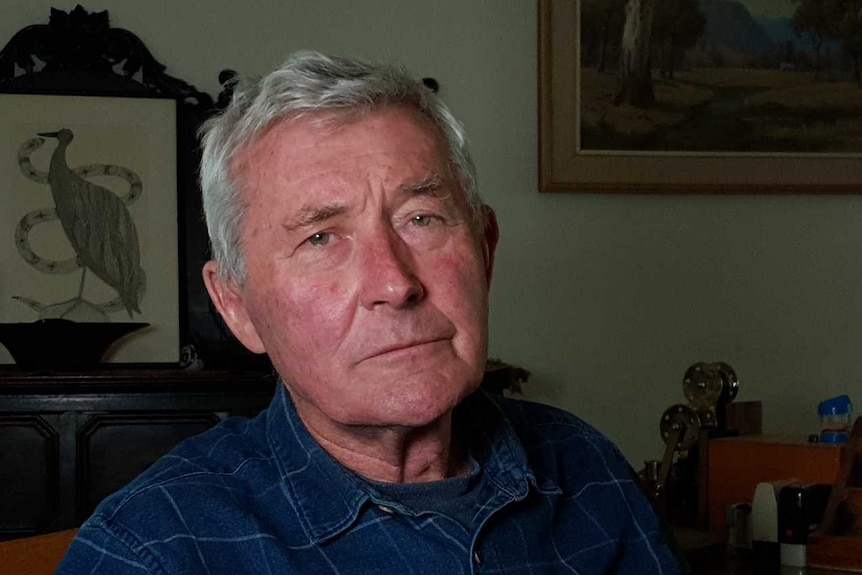 Bernard Collaery wearing a dark blue shirt with light blue stripes, sitting at a wooden desk in his front room office