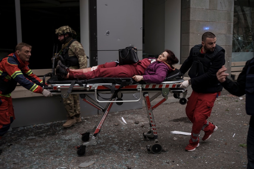 A crying woman with her purse on her lap lies on a stretcher pushed by Red Cross medics 